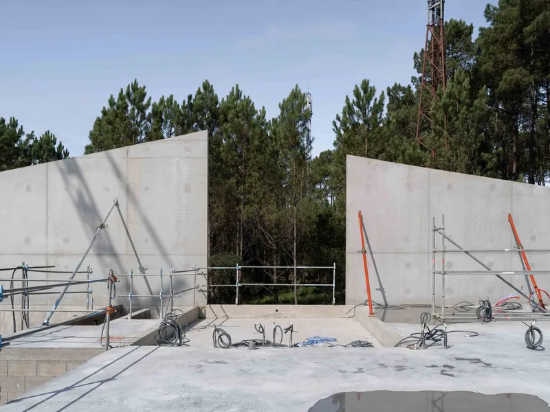 Construction site view showing concrete walls and scaffolding with pine trees in the background