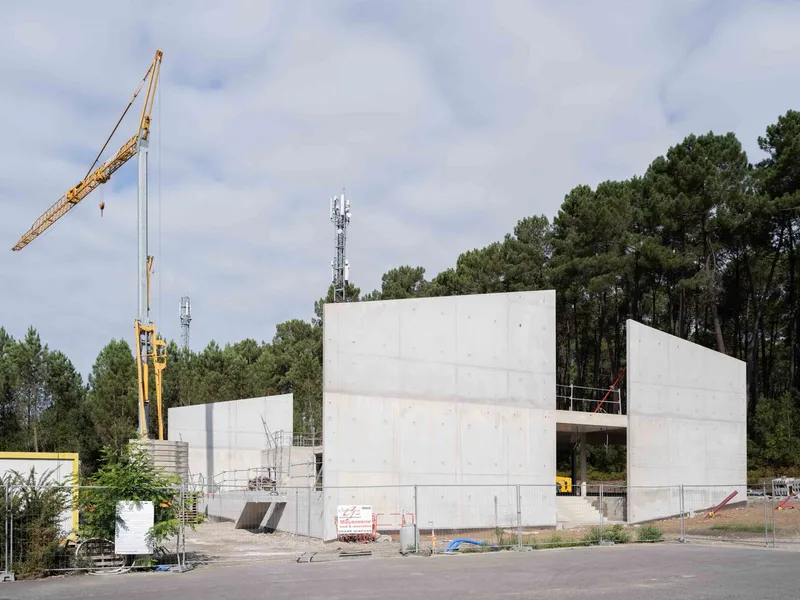 Construction view of concrete volumes with a yellow crane visible against the tree line