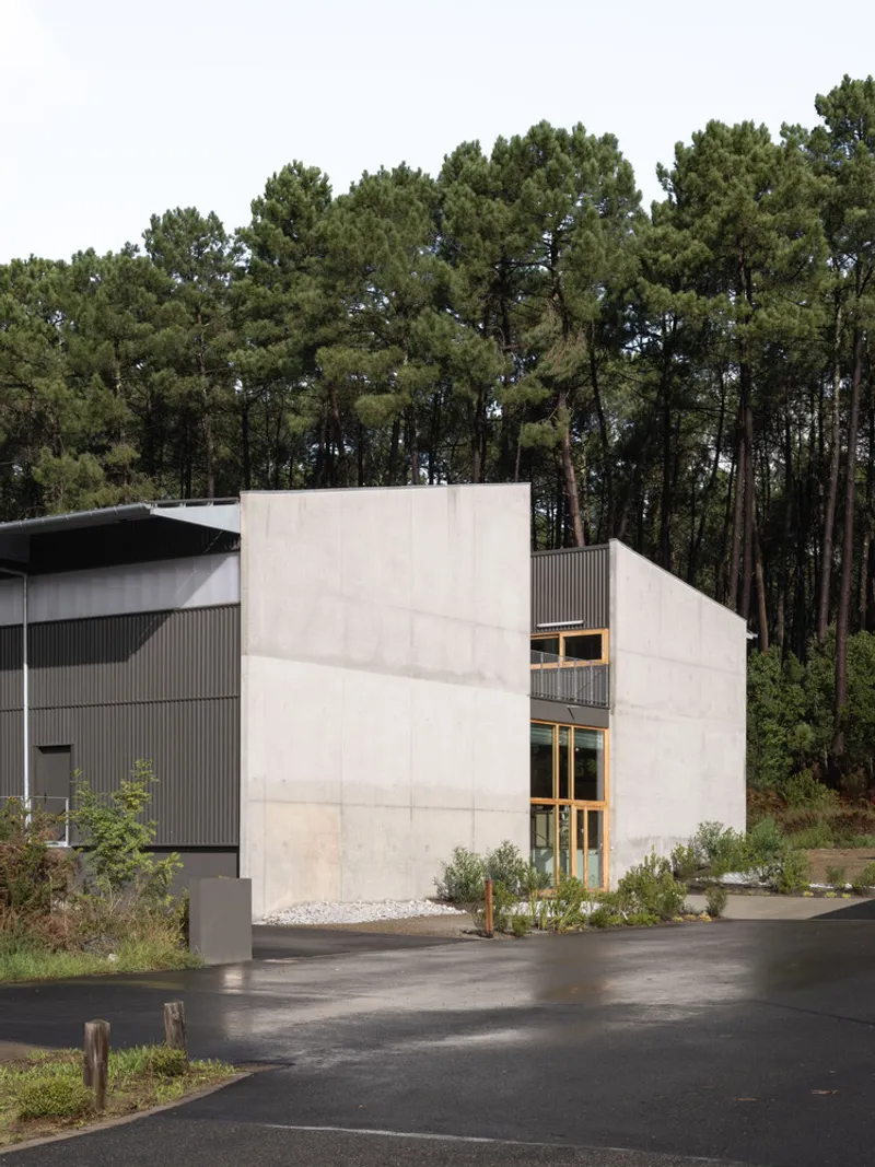 Concrete volume with glazed openings next to a corrugated metal building backed by dense pine forest