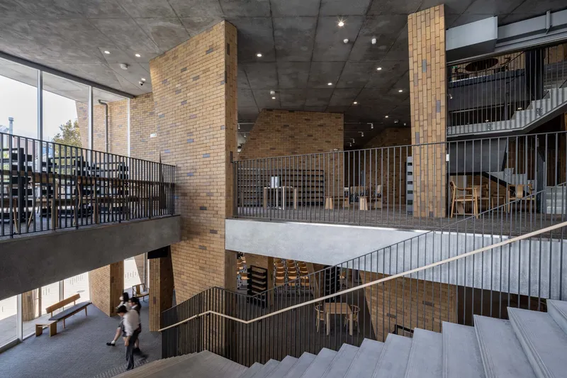 Multi-level atrium interior with exposed concrete ceiling, tan brick columns and people walking across a bridge