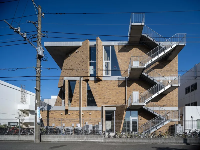 Street view of the tan brick facade with angled structural elements and external steel staircase against a clear blue sky