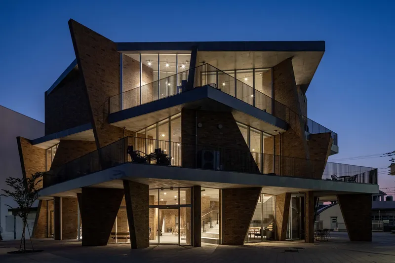 Three-story facade with angled brick volumes and glazed openings illuminated at dusk under a clear sky