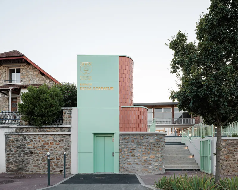 Street entrance: pale green elevator tower with laser-cut signage beside the coral terracotta stair volume, stone wall, trees