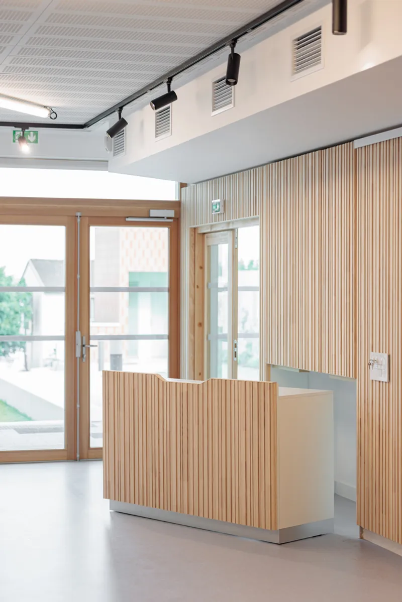 Reception: oak-slatted desk with metal counter, glass entrance doors, track lighting, white walls