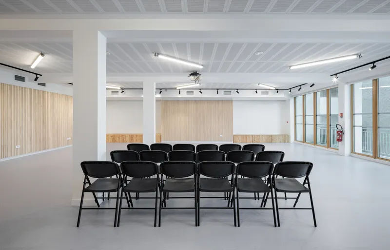 Lecture setup: black folding chairs facing the oak-slatted wall, column, track lighting, daylight through glass doors