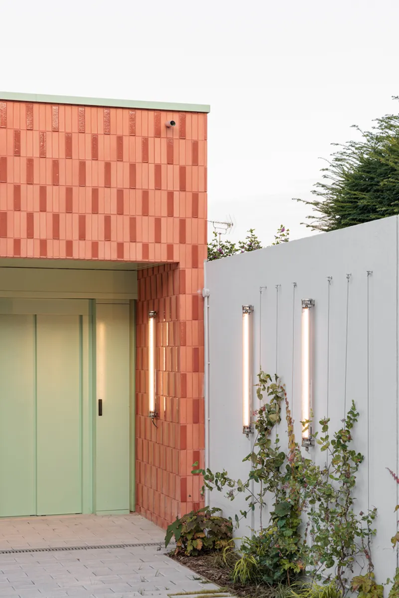 Detail: coral terracotta wall meeting pale green door and white metal screen, vertical slot lights, climbing plants