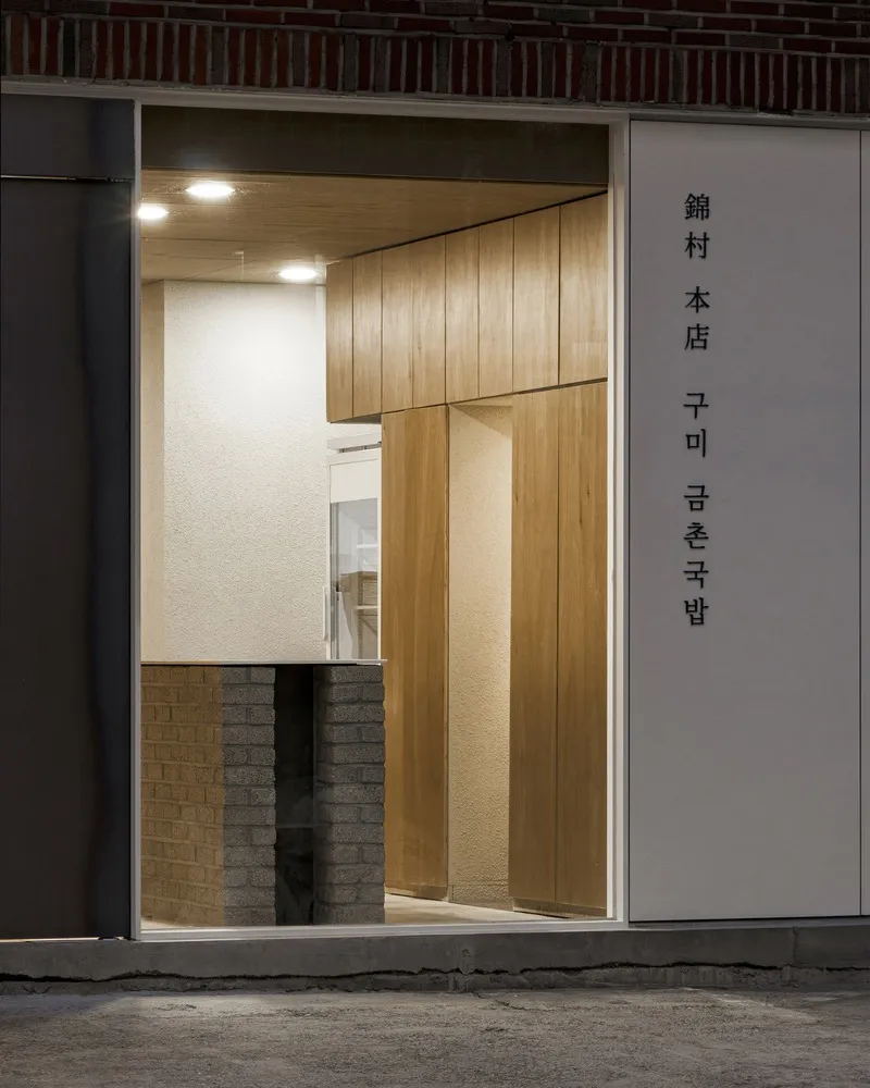 Entrance at night: illuminated doorway framed in oak, white signboard with Korean text, brick surround