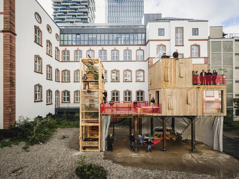 Daytime: two-storey timber and steel structure in a Frankfurt courtyard, steel frame ground floor, timber modules above, red balustrade, people on the terrace