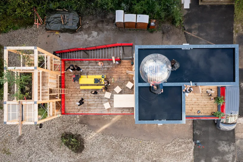 Aerial view: open timber deck with red railing and yellow table at left, dark-roofed residential module with transparent dome at right, gravel ground