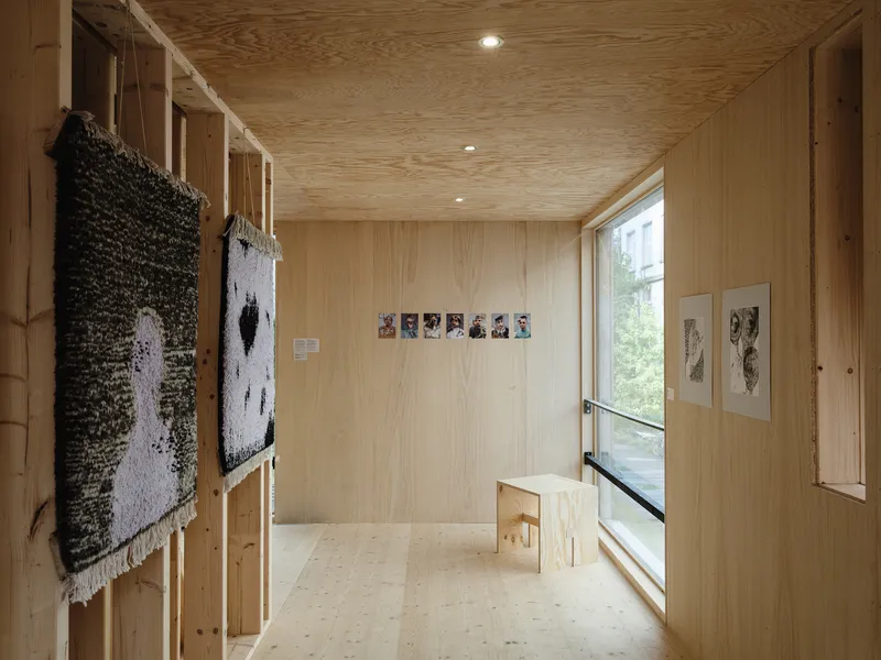 Interior gallery: plywood walls and ceiling, textile artwork hanging, photographs pinned to the wall, plywood stool, narrow window to the courtyard