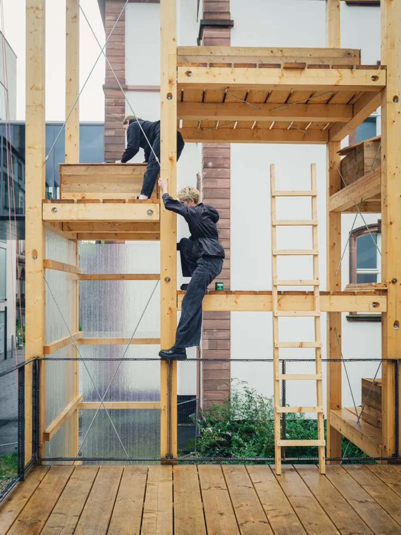 Detail: person climbing a timber ladder between stacked timber-framed modules, polycarbonate panels, plants growing at the edges