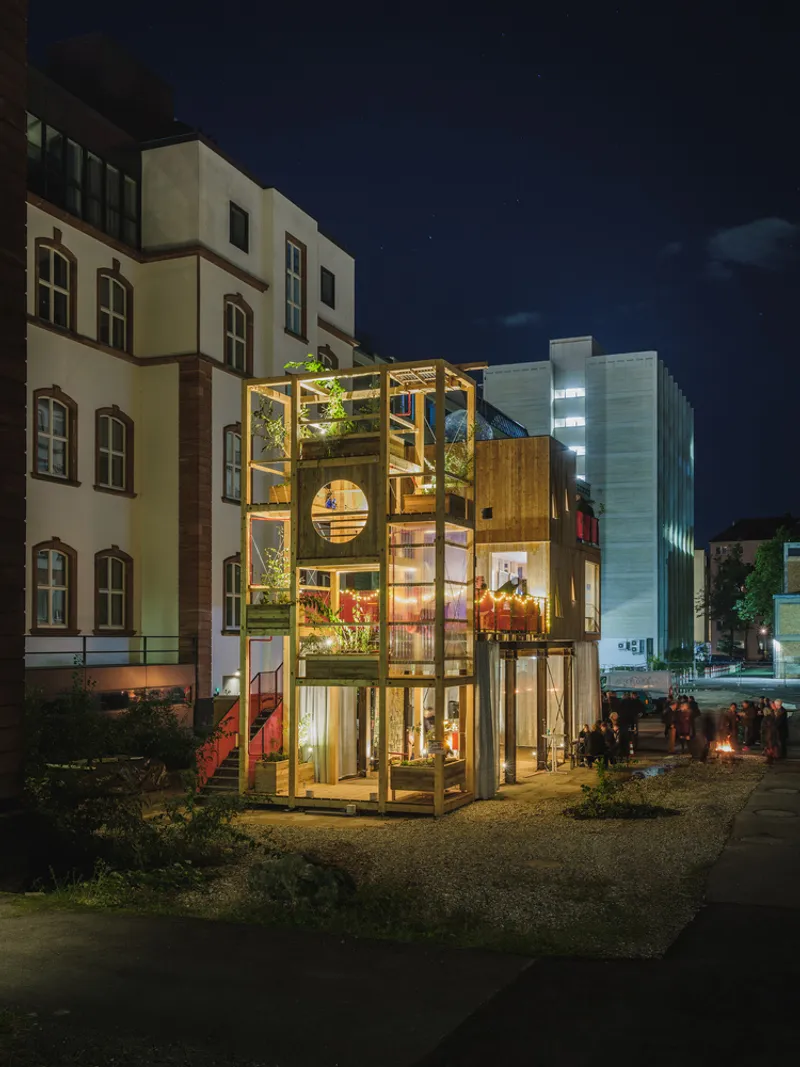 Night: full structure glowing, timber tower with plants at left, timber-clad residential block at right, crowd gathered in the market hall below