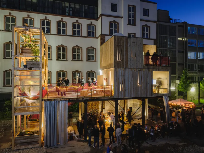 Night wide view: market hall on the ground floor with tables and lights, residential modules above, red balustrade terrace, people, historic courtyard
