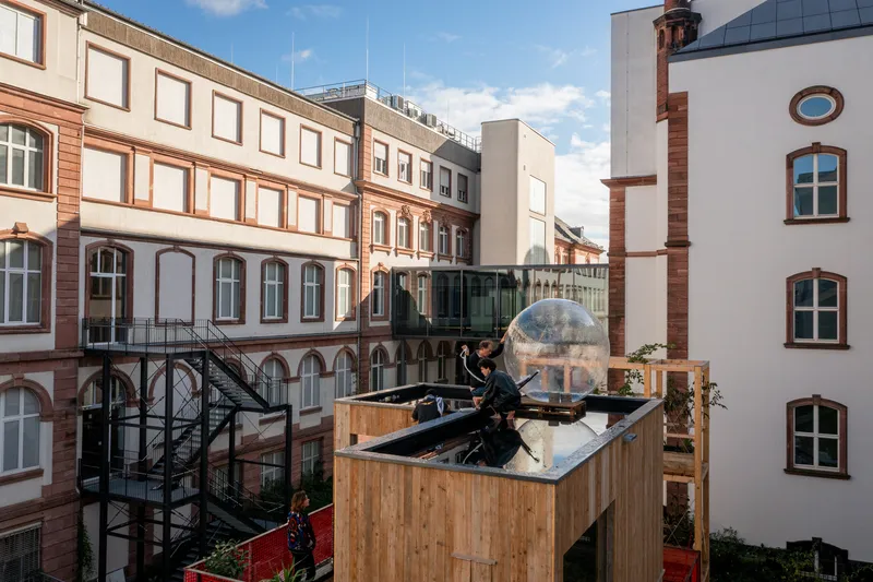 Rooftop: timber-clad residential modules, transparent geodesic dome, chimney structures, historic Frankfurt buildings behind