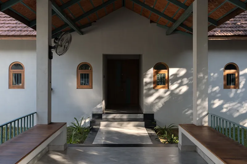 Covered entrance with white walls, arched timber-framed windows, exposed clay roof tiles, and dappled sunlight