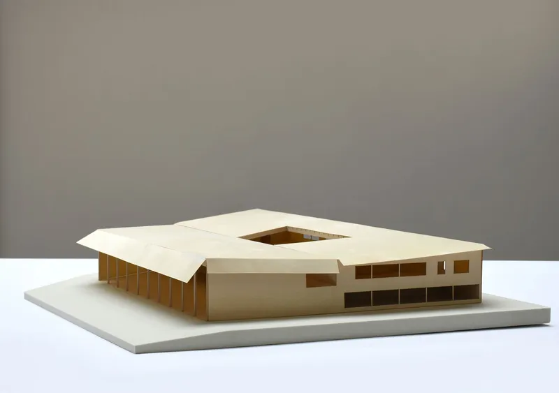 Physical model showing square courtyard void ringed by white roof and bronze colonnade in neutral studio light