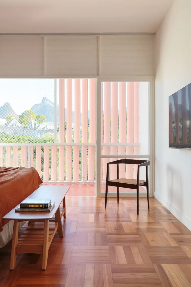 Bedroom window: vertical cobogó screen in pink light, rosewood chair, parquet floor, view to garden and hills beyond