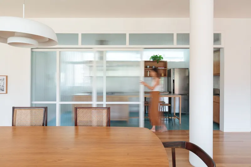 Dining to kitchen: glass sliding partition open, figure moving through, cane chairs, white column, stainless steel cabinets visible