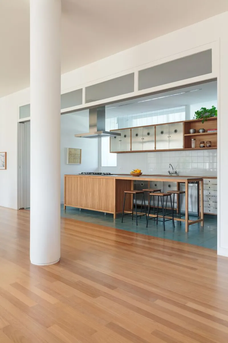 Kitchen from living: white cylindrical column, oak floor meeting blue-green tile, stainless steel island and cabinets