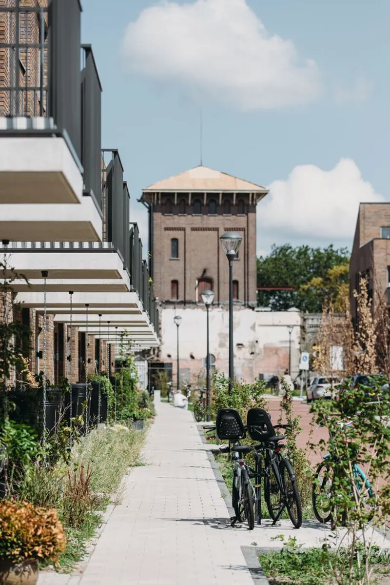 Pedestrian pathway with planted beds and bicycles leading toward a preserved brick water tower under summer clouds