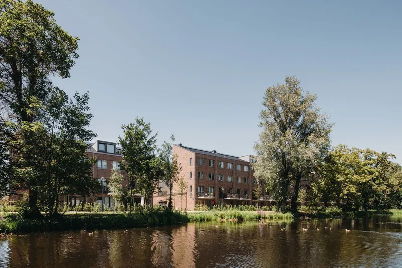 Brick apartment building beyond a waterway lined with dense trees on a clear afternoon
