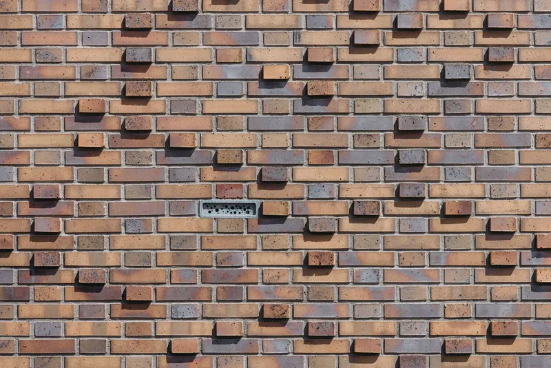 Close-up of variegated brick wall in orange and grey tones with a small ventilation grille in direct sunlight