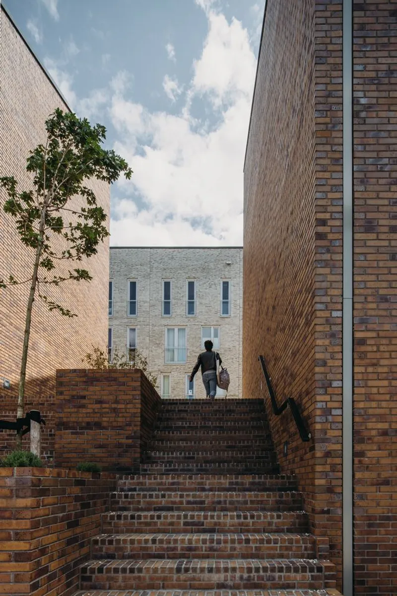 Brick staircase ascending between brick walls as a person walks upward beneath a clouded sky