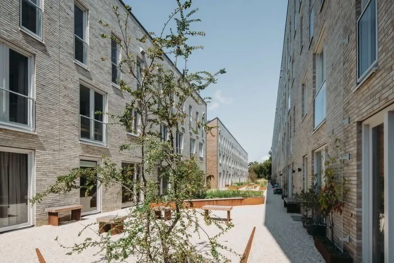 Courtyard between brick buildings with young trees, timber benches and planted beds on gravel