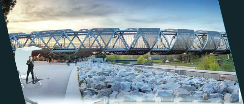 View through an angular concrete frame toward a blue steel truss bridge spanning a rocky channel with a photographer in the foreground