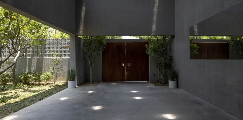 Concrete overhang sheltering timber entry doors with dappled sunlight across the paved forecourt