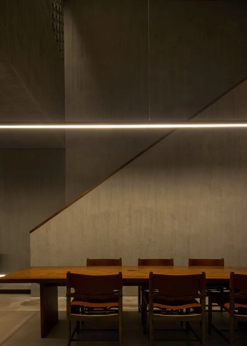 Dining table under recessed linear lighting with concrete walls and wooden staircase