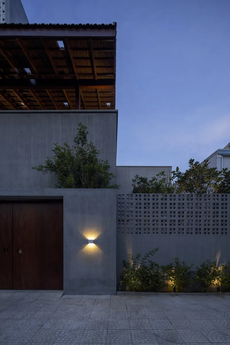 Street facade with perforated breeze block wall and wall-mounted sconce illuminating planted bed at dusk