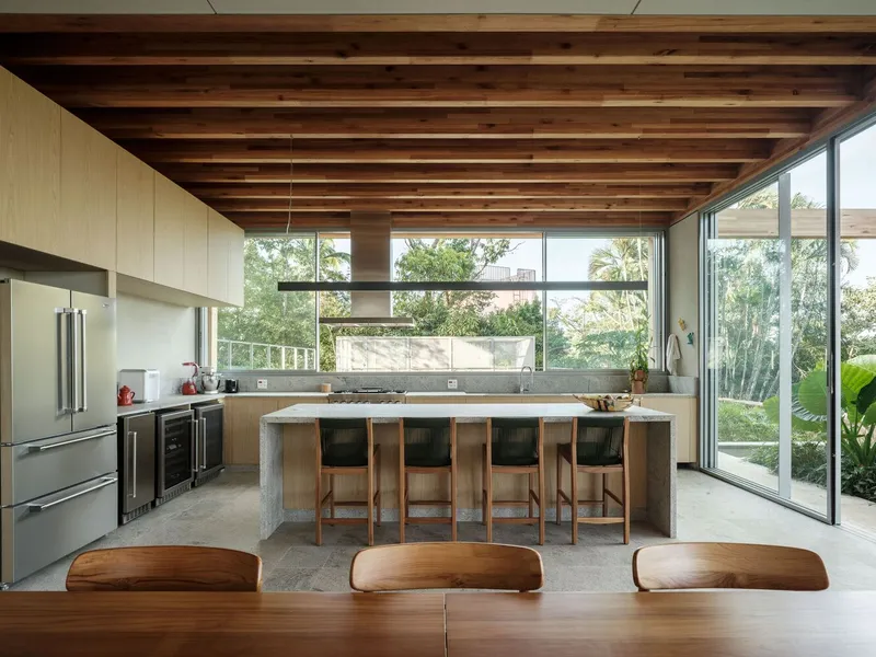 Open kitchen with exposed timber ceiling beams and full-height glazing overlooking garden vegetation