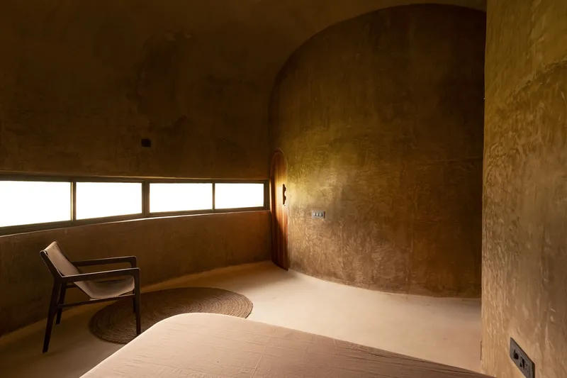 Second bedroom: vaulted earth walls, strip windows catching golden light, round bed, chair, warm monochrome