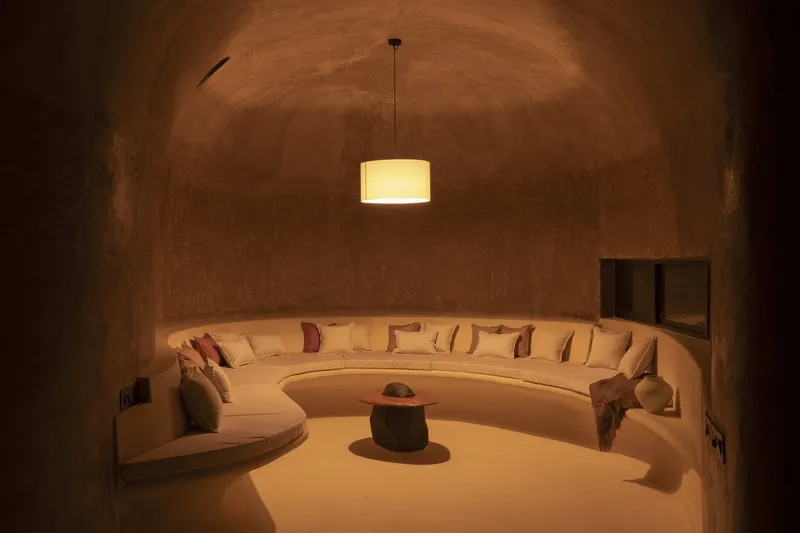 Living room at night: circular sunken sofa, cushions, pendant lamp, vaulted earth ceiling, warm amber glow