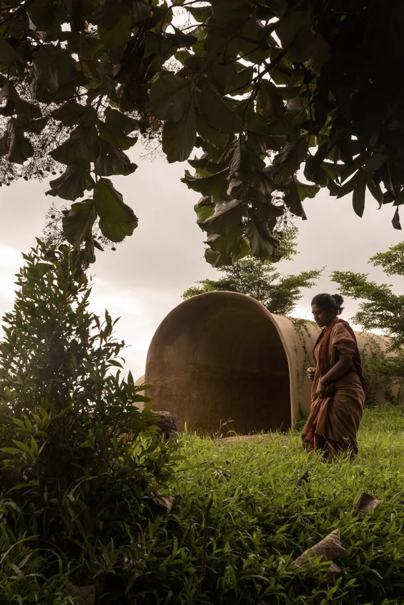 Woman approaching: figure in sari walking toward the arched entry through foliage, earth form behind