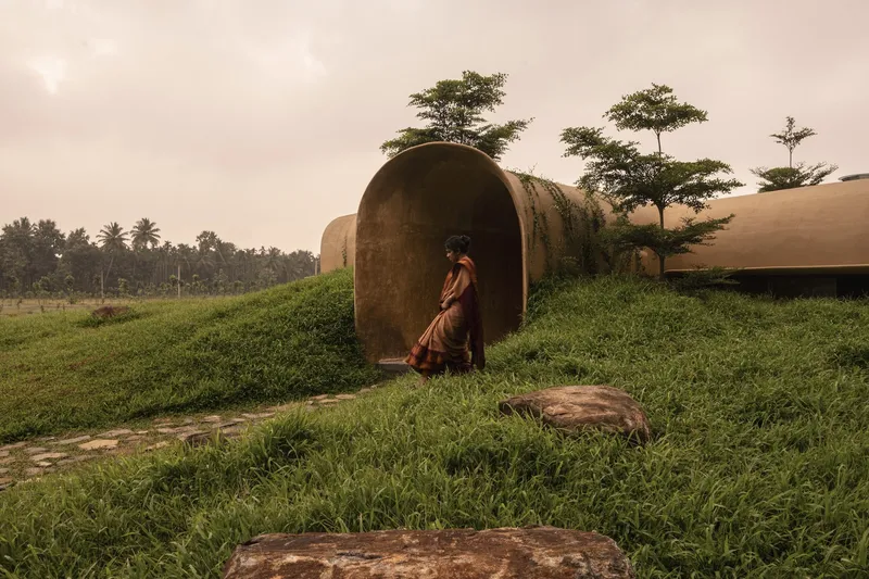 Entry: woman in sari entering an arched earth tunnel, grass-covered mound, palm trees, overcast sky
