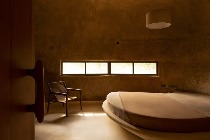 Bedroom: circular earthen bed platform, vaulted ceiling, strip windows, chair, warm earth tones throughout