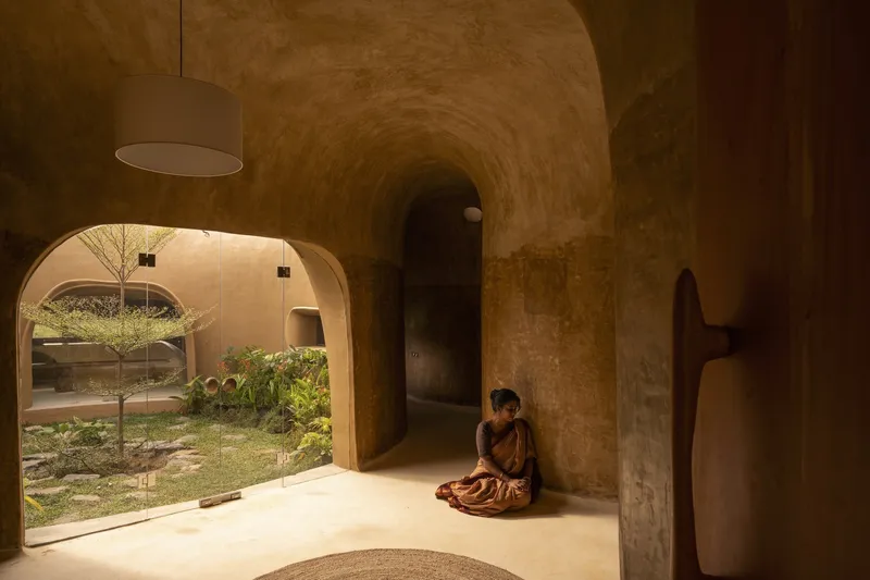 Interior passage: vaulted earth corridor with arched openings, woman seated on the floor, garden visible through doorway