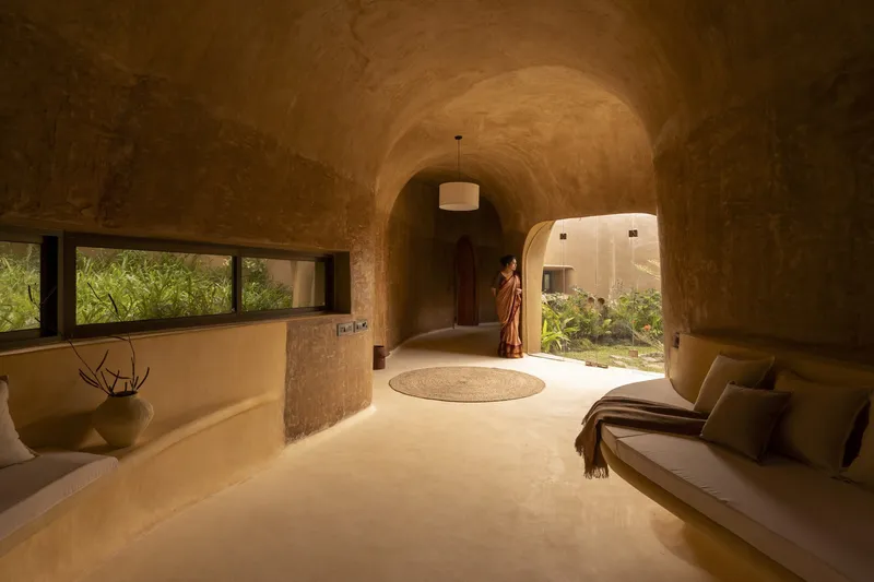 Living room: vaulted earth interior, built-in daybed, arched doorways, circular floor detail, woman in sari, garden view