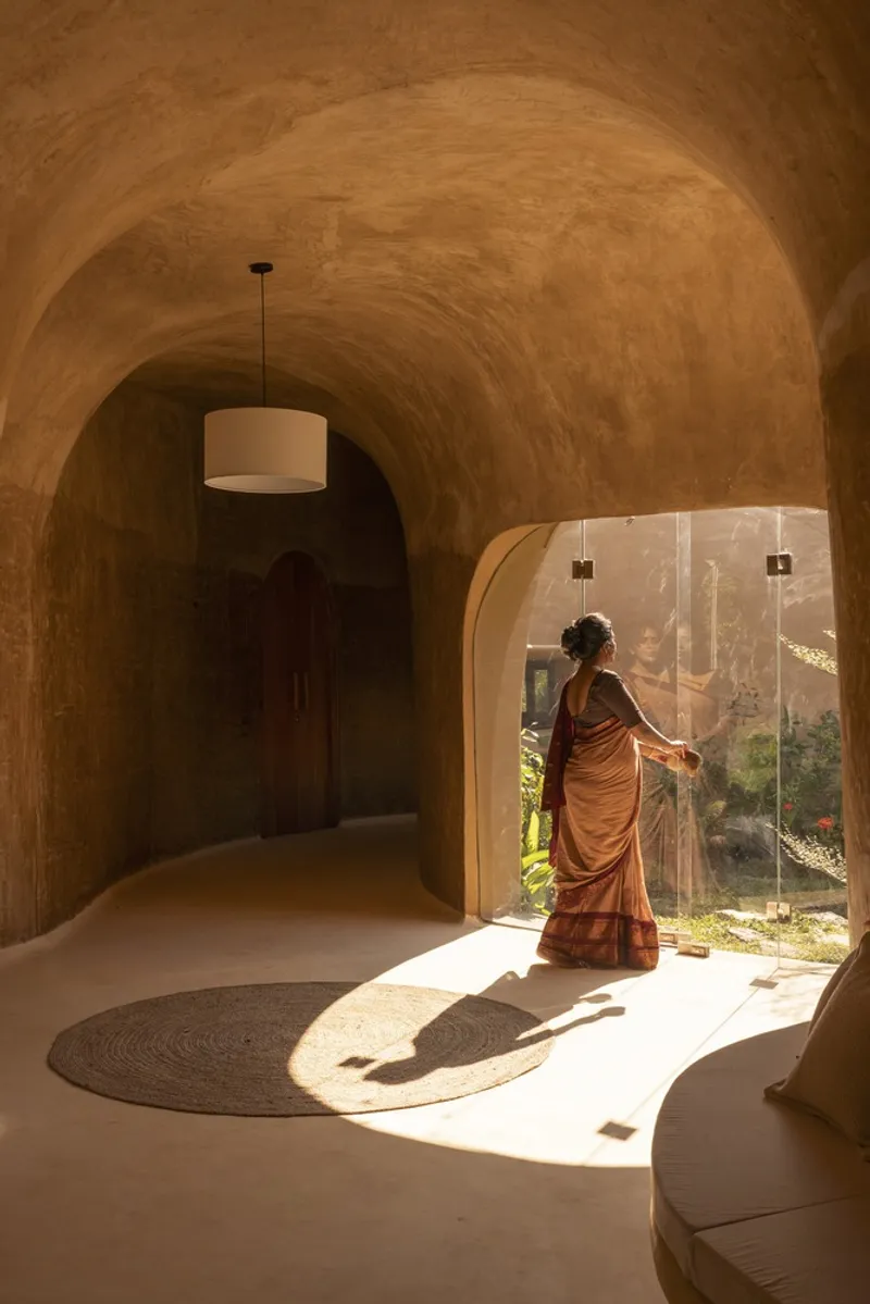 Corridor threshold: woman silhouetted in arched doorway, sun casting a circle on the floor, earth vault above