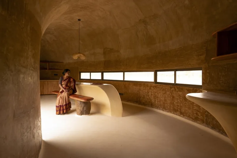 Kitchen with woman: barrel vault, built-in earthen island and counter, strip windows, pendant lamp, lime plaster