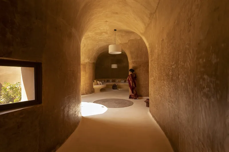 Corridor perspective: vaulted earth passage leading to sunken living room, woman seated at the far end, pendant light