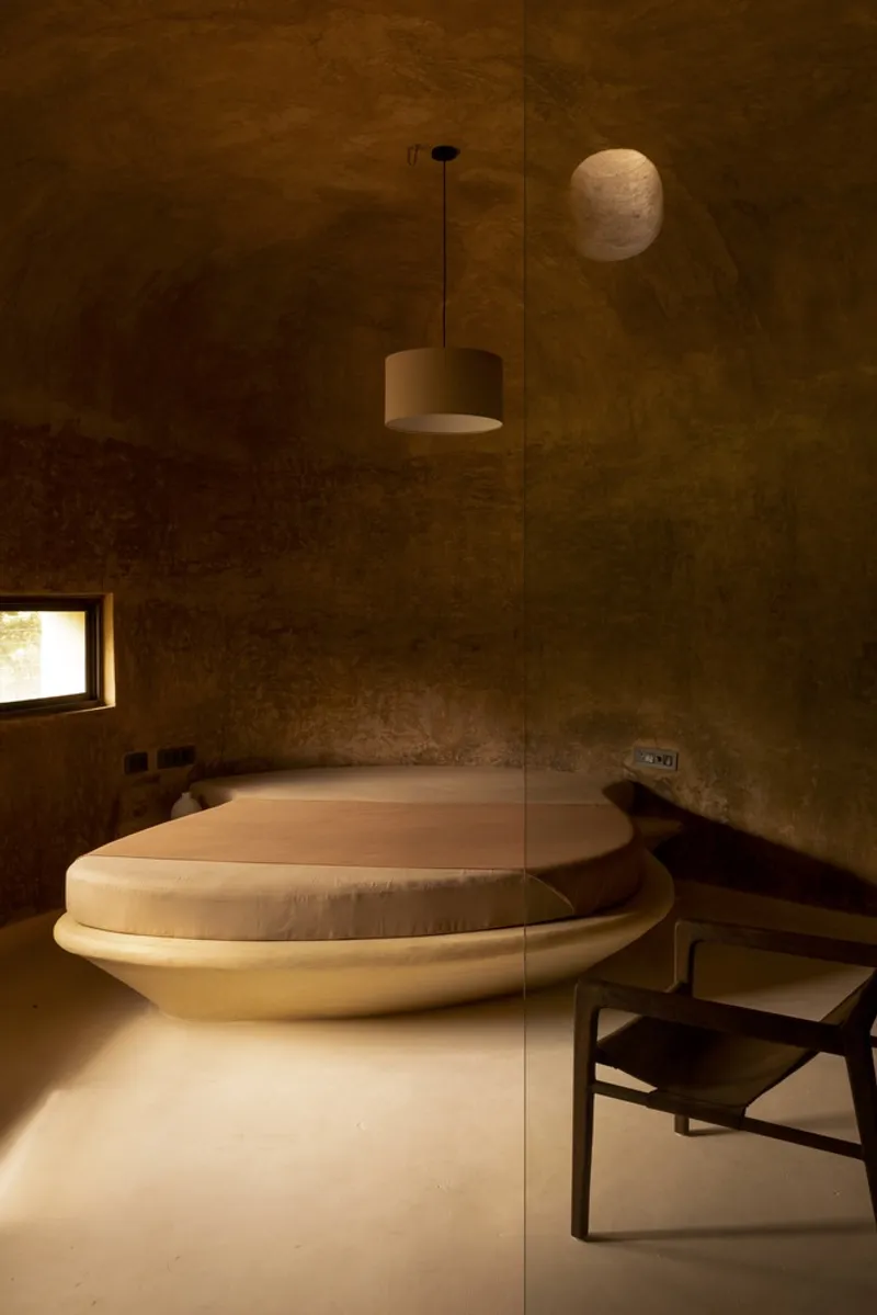 Bedroom detail: circular bed platform, vaulted earth ceiling, pendant lamp, warm amber light, chair