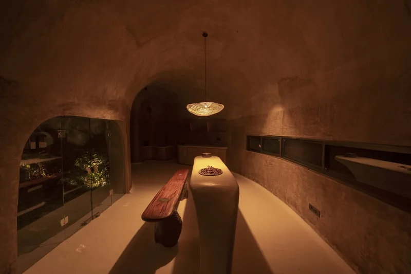 Dining at night: vaulted earth room, long built-in counter with timber top, pendant lamp, arched garden window