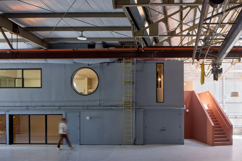 Grey steel volume with circular window and yellow ladder inside converted industrial hall with exposed trusses