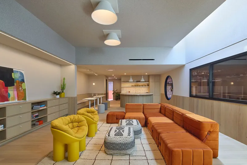 Communal lounge with orange modular seating and yellow sculptural chairs beneath blue-grey ceiling