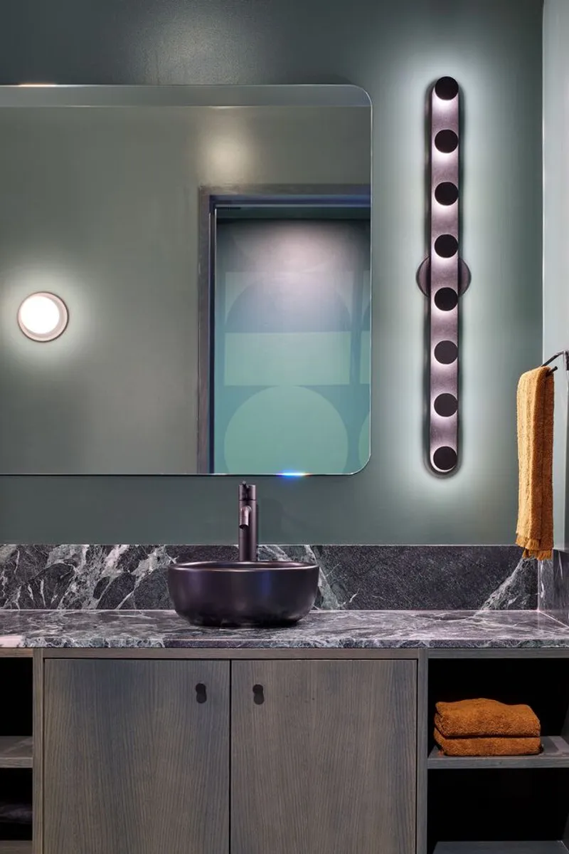 Bathroom vanity with dark stone counter, vessel sink, backlit mirror, and vertical tubular sconce