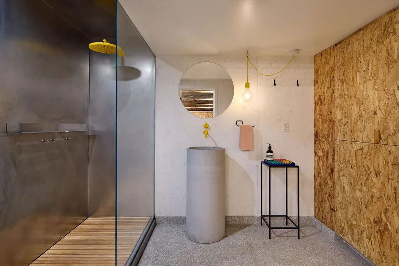 Bathroom with cylindrical concrete sink, circular mirror, oriented strand board wall, and glass-enclosed shower