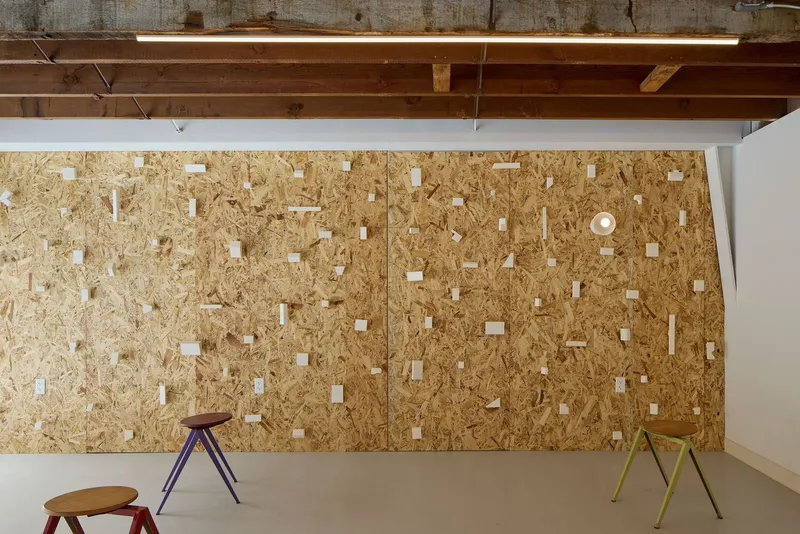 Oriented strand board wall with exposed timber ceiling beams and scattered wooden mounting blocks under linear fluorescent lighting